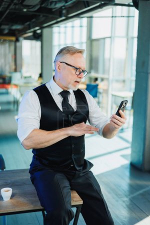 Téléchargez les photos : Un homme d'affaires barbu aux cheveux gris tient un téléphone dans ses mains, un homme regarde une vidéo au téléphone. Travailler au bureau - en image libre de droit