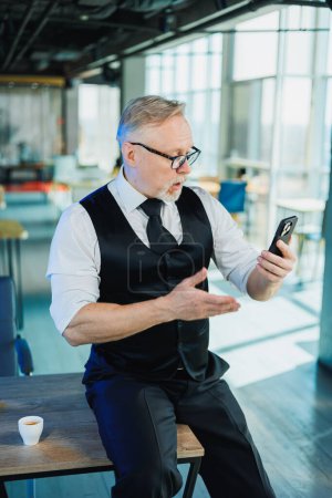 Téléchargez les photos : Un homme d'affaires barbu aux cheveux gris tient un téléphone dans ses mains, un homme regarde une vidéo au téléphone. Travailler au bureau - en image libre de droit