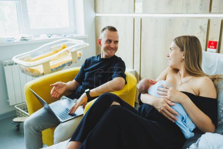 Der junge Vater arbeitet online am Laptop, während seine Frau mit dem Baby spielt. Junges glückliches modernes Paar.