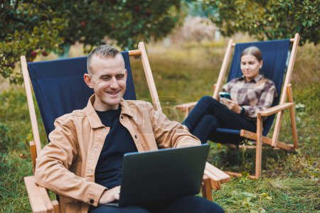 Konzentrierter Mann im Garten, der an einem Laptop arbeitet, während er auf einem Stuhl zwischen Bäumen sitzt. Ein Mann arbeitet fernab vom Garten und denkt über eine neue Geschäftsidee oder kreative Idee nach.