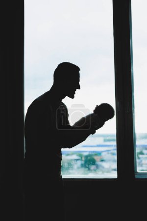 Photo for A young loving dad holds his son in his arms against the background of the window. - Royalty Free Image