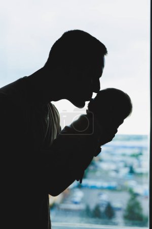 Photo for A young loving dad holds his son in his arms against the background of the window. - Royalty Free Image