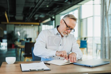 Ein grauhaariger Geschäftsmann mit Brille sitzt an einem Schreibtisch und arbeitet mit Papieren.