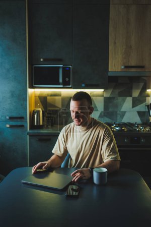 A young man sits at the table in the kitchen and works on a laptop. The laptop is on the table and the young man is working at home.