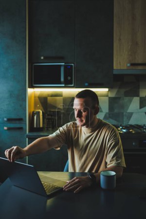 A young man sits at the table in the kitchen and works on a laptop. The laptop is on the table and the young man is working at home.