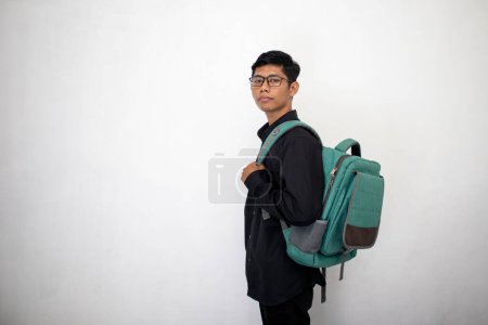Téléchargez les photos : Homme asiatique en vêtements noirs soignés porte un sac à dos vert sur un fond blanc. un étudiant portant un sac en toute confiance. - en image libre de droit