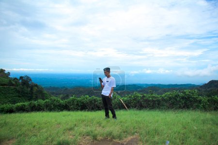 Photo for Asian man using phone outdoors. Young man standing in green meadow under cloudy sky - Royalty Free Image