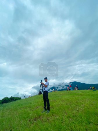 Téléchargez les photos : Asiatique homme avec caméra debout à prairie verte, ciel nuageux arrière-plan - en image libre de droit