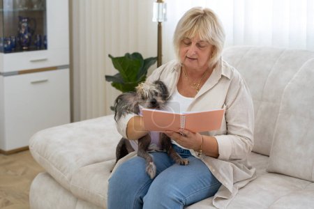Photo for Older caucasian woman writing notes in notebook planner, sitting with dog on sofa in living room at home. - Royalty Free Image