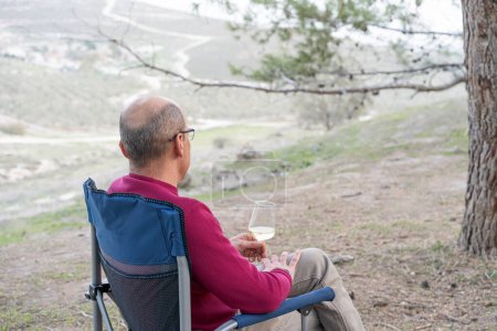 Photo for Man sitting on folding chair with glass of wine, looking forward at nature. Back view. - Royalty Free Image