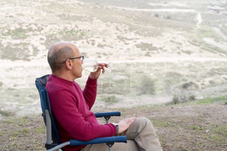 Photo for Man sitting on folding chair, drinking wine, relaxing and admiring the beautiful views of nature. - Royalty Free Image