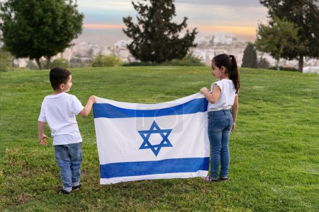 Photo for Young children stand on a hill holding an Israeli flag. A rear view little girl and boy with an Israel flag in the sunset. - Royalty Free Image