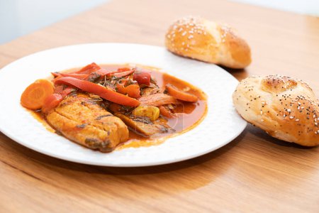 Delicious Jewish Shabbat Delight: Moroccan Fish Chraime with Vibrant Tomato Sauce, Peppers, and Carrots, served on a White Plate. Side of Challah Buns. Traditional, Festive, Tasty.