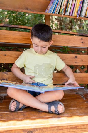 Photo for Young Child Absorbed in Books at Street Library on a Wooden Bench. Boy Immersed in Summer Reading on Wooden Bench. - Royalty Free Image