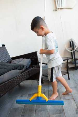 Photo for Funny young child cleaning floor at home. Boy washing floor with floor squeegee in balcony of modern apartment. Male household helping tidy house. - Royalty Free Image