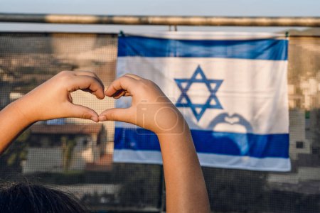 Zärtliche Kinderhände schaffen eine Herzform, Sonnenlicht wirft Herzschatten auf die israelische Nationalflagge, die auf dem Balkon hängt.