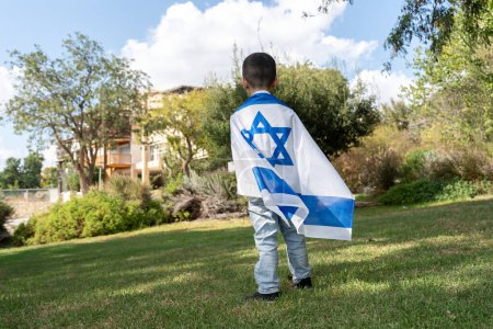 Niño envuelto en una bandera israelí se para y mira un asentamiento israelí.