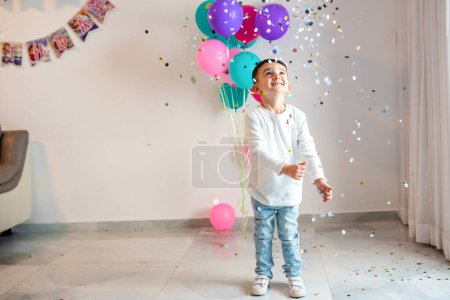 A delighted young boy with a bunch of colorful balloons and flying confetti celebrates at his birthday party, full of smiles and excitement.