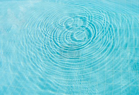 Klares blaues Poolwasser mit Wellen, die nach außen strahlen und ein faszinierendes Muster auf dem gefliesten Beckenboden erzeugen. Bild fängt die ruhige und erfrischende Essenz eines ruhigen, sonnigen Tages am Pool ein.