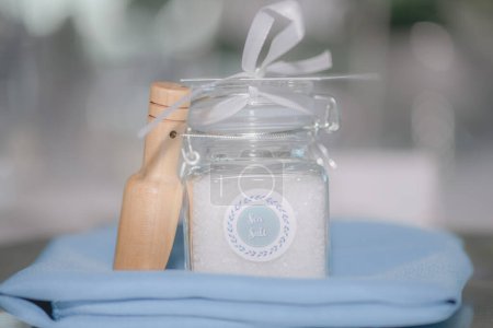 Téléchargez les photos : Belle mer sel rempli pot clair avec arc blanc mariage faveur avec du bois sur couverture bleue en face de bokeh blanc - en image libre de droit