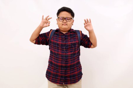 Téléchargez les photos : Détendu paisible asiatique écolier garçon méditant avec les yeux fermés. Garder les mains dans le geste Mudra, debout isolé sur fond blanc - en image libre de droit