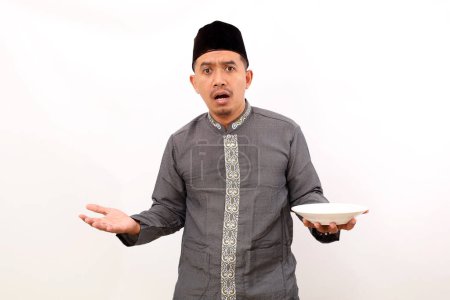 Photo for Unhappy asian muslim man standing while holding an empty plate. Isolated on white background - Royalty Free Image