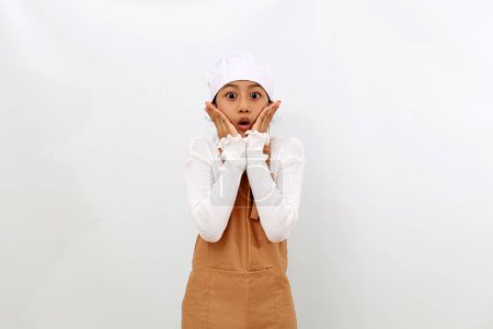 Photo for Shocked asian little girl in chef uniform standing while looking at the camera. Isolated on white background - Royalty Free Image