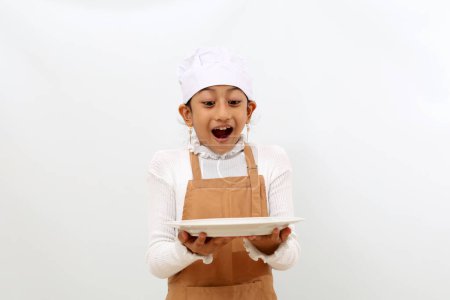 Foto de Wow and surprised expression of asian little girl in chef uniform holding empty plate. Isolated on white background - Imagen libre de derechos
