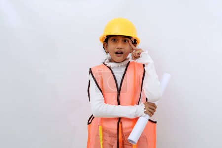 Photo for Cute Asian little girl in the construction helmet as an engineer holding a blueprint paper while pointing her head. Isolated on white - Royalty Free Image