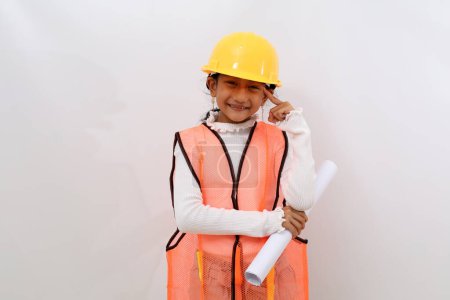 Photo for Happy Asian little girl in the construction helmet as an engineer holding a blueprint paper while pointing her head. Isolated on white - Royalty Free Image