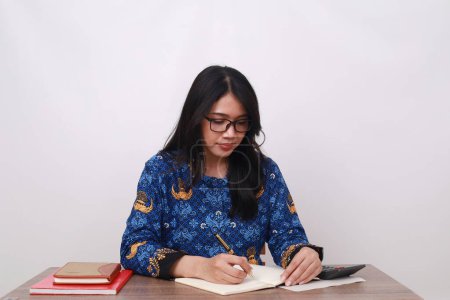 Foto de Mujer asiática en batik korpri, uniforme obrero tradicional indonesio escribiendo algo en un libro - Imagen libre de derechos