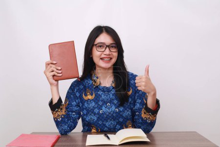 Téléchargez les photos : Femme asiatique en batik korpri, indonésien traditionnel ouvrier uniforme holding book, montrant pouces vers le haut - en image libre de droit
