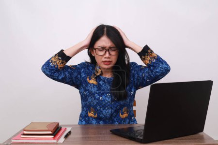 Téléchargez les photos : Stressé asiatique femelle en batik korpri, indonésien traditionnel uniforme tenant sa tête tout en travaillant - en image libre de droit