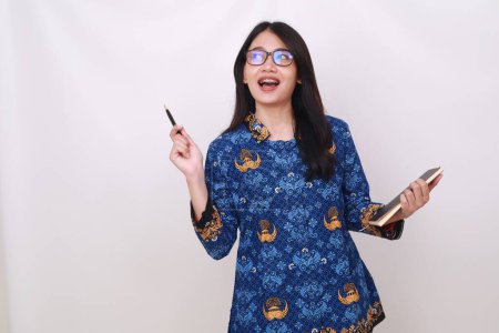 Téléchargez les photos : Femme asiatique en batik korpri, uniforme indonésien traditionnel tenant un livre et pointant latéralement - en image libre de droit