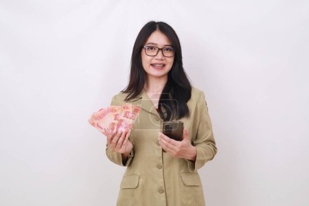 Photo for Young pretty teacher holding Indonesian banknotes and cell phone while looking at the camera - Royalty Free Image