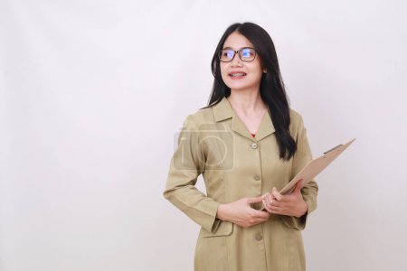 Photo for Pretty young Indonesian teacher standing while holding clipboard and looking sideways on empty space - Royalty Free Image