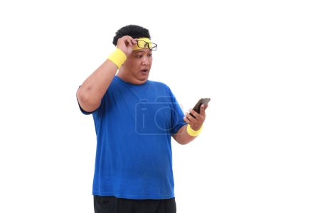 Photo for Wow shocked Asian overweight man in sportswear standing while holding a cell phone. Isolated - Royalty Free Image