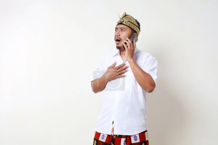 Photo for Shocked Asian man in balinese traditional costume standing while talking on the phone and looking sideways. Isolated on white background - Royalty Free Image