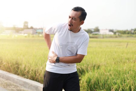 Photo for Asian man with her muscle pain during running. Runner man having back and waist body ache due to Piriformis Syndrome, Low Back Pain and Spinal Compression. Sports injuries and medical concept - Royalty Free Image