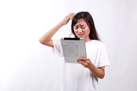 Attraktive junge asiatische Frau hält ein digitales Tablet in der Hand, während sie vor weißem Hintergrund mit verwirrtem Gesichtsausdruck mit dem Kopf kratzt