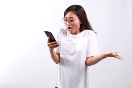 Shocked asian business woman asking and shouting at mobile phone, looks outraged, furious while reading something on smartphone, standing over white background