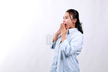 Schockiert und erschrocken blickt die asiatische Frau zur Seite in den leeren Raum, bedeckt den Mund mit den Händen sprachlos, steht vor weißem Hintergrund.