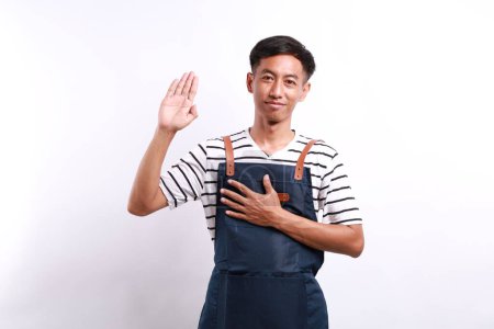 Asiatischer junger Mann mit blauer Schürze über weißem Hintergrund schwört mit der Hand auf der Brust und offener Handfläche und leistet einen Treueschwur