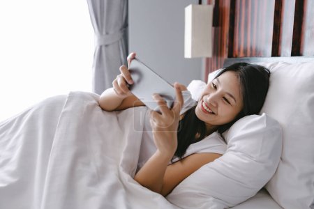 Téléchargez les photos : Belle femme asiatique heureuse en utilisant téléphone mobile sur le lit après le réveil dans la chambre moderne à la maison - en image libre de droit