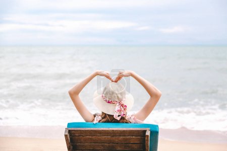 Téléchargez les photos : Voyage d'été concept de vacances à la plage, Heureux voyageur asiatique femme avec chapeau se détendre et faire forme de coeur sur la plage de la chaise à Pattaya, Chon Buri, Thaïlande - en image libre de droit