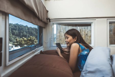 Téléchargez les photos : Voyage vacances en camping-car concept, Heureux voyageur asiatique femme avec téléphone mobile se détendre à l'intérieur d'un camping-car véhicule récréatif dans la chambre à coucher à Mon Jam, Chiang Mai, Thaïlande - en image libre de droit