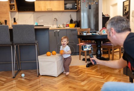 Photo for Adorable one year old child playing with orange while father is recording a video - Royalty Free Image