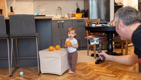 Téléchargez les photos : Adorable enfant d'un an jouant avec de l'orange pendant que le père enregistre une vidéo - en image libre de droit