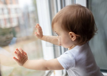 Téléchargez les photos : Portrait d'un adorable enfant d'un an regardant à travers la fenêtre, faible profondeur de champ - en image libre de droit