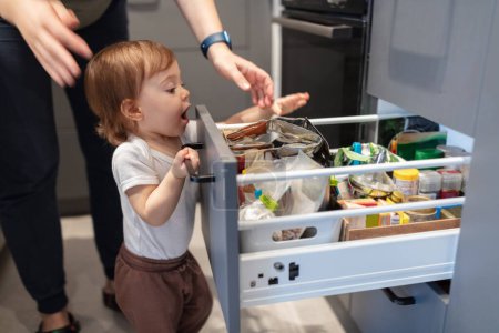 Téléchargez les photos : Portrait d'adorable enfant d'un an regardant à travers le tiroir de la cuisine, faible profondeur de champ - en image libre de droit